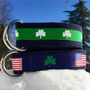 May include: Two navy blue belts with silver metal rings. One belt has a green stripe with white shamrocks, the other has a green shamrock and an American flag. The belts are resting on snow.