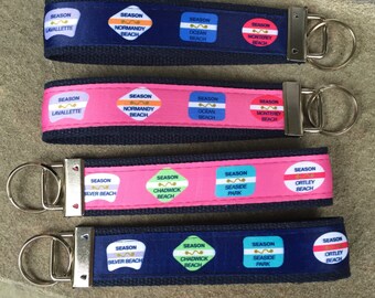 Op de afbeelding: Vier sleutelhangers met verschillende gekleurde stoffen banden en metalen sleutelringen. Elke band heeft een herhalend patroon van kleurrijke vierkanten met de woorden "SEASON" en de naam van een strand of park. De kleuren van de banden zijn blauw, roze, roze en blauw.