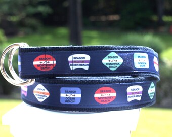 Op de afbeelding: Een donkerblauwe stoffen riem met een herhalend patroon van kleurrijke strandpatches met tekst waaronder Season Monterey Beach, Silver Beach, Chadwick Beach, Seaside Park, Normandy Beach en Ocean Beach, afgewerkt met twee zilverkleurige metalen D-ringen.