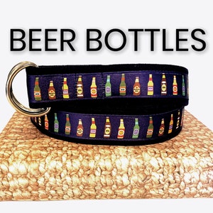 May include: A black fabric belt with a gold buckle, featuring a repeating pattern of colorful beer bottles. The belt is on a woven brown surface. The text "BEER BOTTLES" is displayed above the belt.