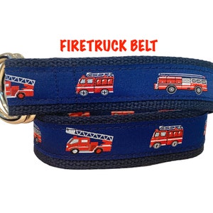 May include: A blue and red fire truck belt with a silver buckle. The belt is made of a woven fabric and features a repeating pattern of fire trucks.