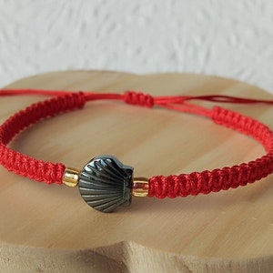 Peut inclure: Un bracelet en cordon rouge doté d'une perle centrale en hématite gris foncé en forme de coquillage, encadrée par deux petites perles métalliques dorées. Le bracelet présente un tressage en macramé et des extrémités coulissantes réglables.