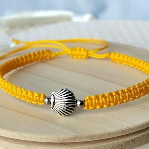 May include: A bright yellow braided cord bracelet featuring a central silver-toned metal seashell charm. The bracelet includes two small silver-toned metal beads flanking the shell and an adjustable sliding knot closure with long cord ends.