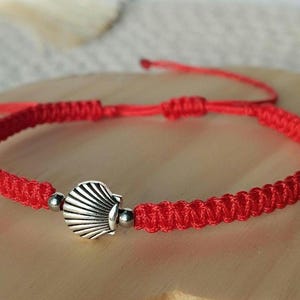 Peut inclure: Un bracelet en cordon tressé rouge avec une breloque centrale en forme de coquillage en métal argenté. Le bracelet est composé d'un cordon tissé et comprend deux petites perles en métal argenté de chaque côté du coquillage, avec des liens réglables.