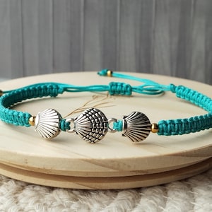 May include: A turquoise cord bracelet featuring three silver-toned metal seashell charms arranged in the center. The bracelet is constructed with a woven macrame knot pattern and includes small gold-toned metal beads flanking the shell charms.