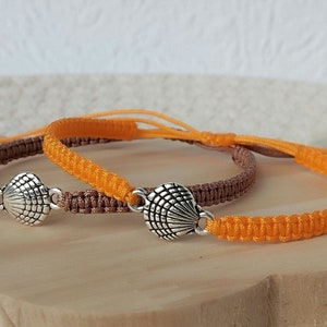 Può includere: Due braccialetti in macramè, uno marrone e l'altro arancione. Ogni braccialetto presenta un ciondolo a forma di conchiglia argentata. I braccialetti sono regolabili con nodi scorrevoli. Sfondo in legno chiaro.