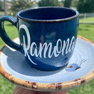 May include: A blue ceramic coffee mug with a white inscription that reads "Ramona" and a matching saucer with a blue and white butterfly design.