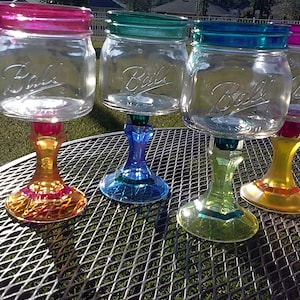 May include: Four glass mason jars with colorful painted rims and bases, transformed into wine glasses. The jars are clear and have the word "Ball" printed on them. The bases are painted in shades of yellow, green, blue, and orange.