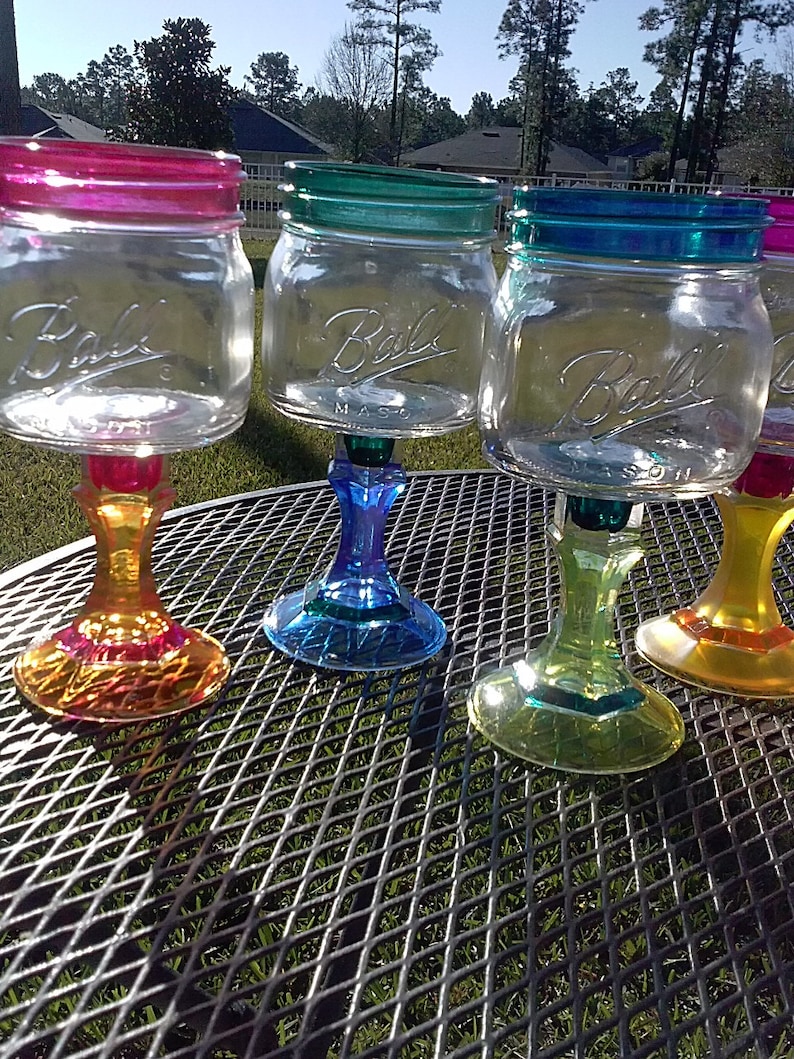May include: Four glass mason jars with colorful painted rims and bases, transformed into wine glasses. The jars are clear and have the word "Ball" printed on them. The bases are painted in shades of yellow, green, blue, and orange.