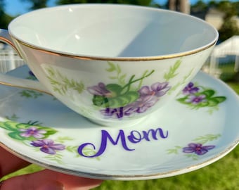 Peut inclure: Une tasse à thé et une soucoupe blanches avec une bordure dorée et un motif floral de violettes violettes. La soucoupe porte le mot "Maman" écrit en cursive violette.