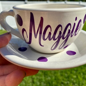 May include: White teacup and saucer with purple polka dots. The teacup has the name "Maggie" written in purple cursive.
