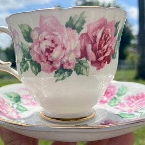 May include: A white teacup and saucer set with a pink rose floral pattern. The teacup has a gold rim and a delicate handle. The saucer has a matching floral design and a gold rim.