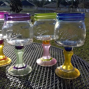 May include: Four glass mason jars with colorful painted bases and rims, repurposed as wine glasses. The jars are clear glass with the word "Ball" printed on the side. The bases are painted in shades of purple, green, pink, and yellow.