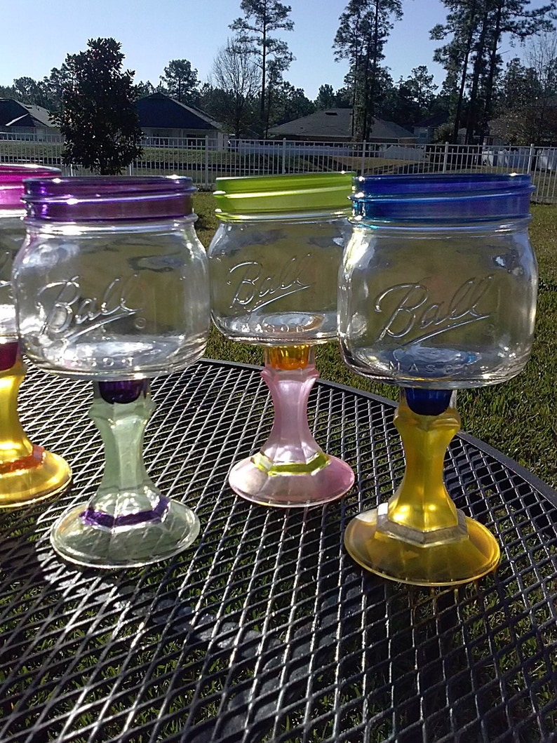 May include: Four glass mason jars with colorful painted bases and rims, repurposed as wine glasses. The jars are clear glass with the word "Ball" printed on the side. The bases are painted in shades of purple, green, pink, and yellow.