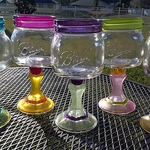 May include: Four glass mason jars with colorful painted bases and rims, transformed into wine glasses. The jars are clear glass with the words "Ball" printed on the front. The bases are painted in shades of yellow, green, pink, and purple.