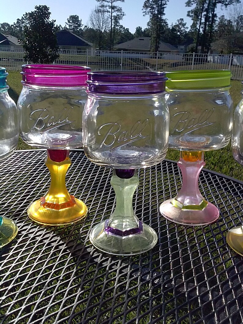 May include: Four glass mason jars with colorful painted bases and rims, transformed into wine glasses. The jars are clear glass with the words "Ball" printed on the front. The bases are painted in shades of yellow, green, pink, and purple.