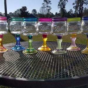 May include: Eight clear glass mason jars with the word "Ball" embossed on the side, painted in various colors and attached to colorful glass stems, arranged on a black metal table.