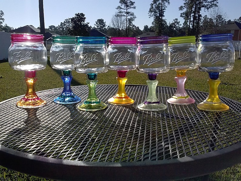 May include: Eight clear glass mason jars with the word "Ball" embossed on the side, painted in various colors and attached to colorful glass stems, arranged on a black metal table.