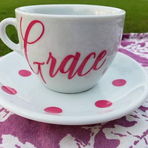 May include: A white teacup and saucer with pink polka dots. The teacup has the word "Grace" written in pink cursive on the side.