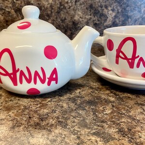 May include: White teapot and teacup set with pink polka dots and the name "Anna" written in pink on both pieces.