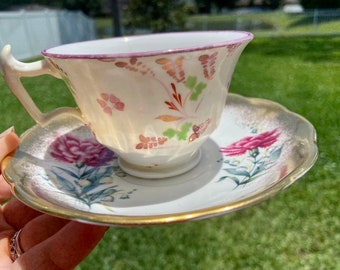 May include: A vintage teacup and saucer set with a floral design. The cup and saucer are white with pink, green, and gold accents. The saucer has a scalloped edge.