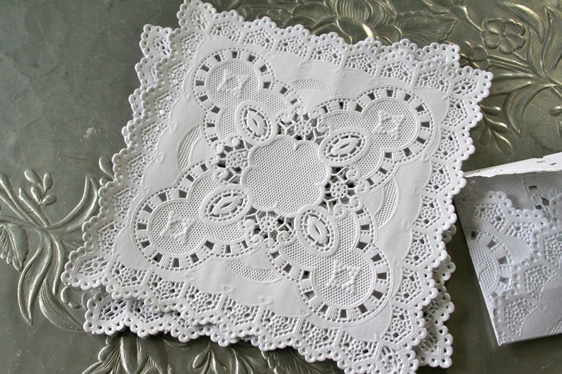 May include: White paper doilies with a delicate lace pattern. The doilies are square shaped and have a scalloped edge.