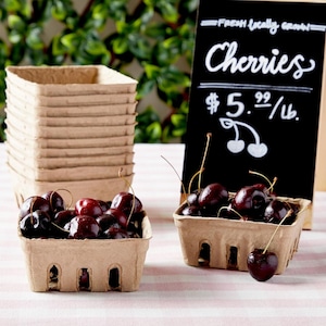 May include: A close-up of two brown cardboard containers filled with fresh, dark red cherries. The containers are on a pink and white striped surface. A chalkboard sign behind the containers reads "Fresh Locally Grown Cherries $5.99/lb."