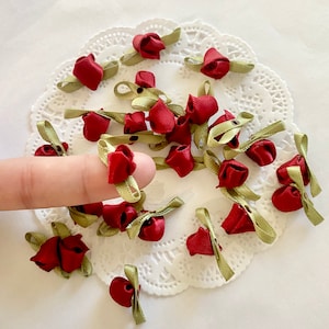 May include: A collection of red and green fabric rose embellishments with a satin finish. The roses are arranged on a white doily.