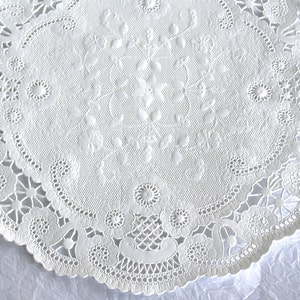 K&ouml;nnte beinhalten: Wei&szlig;e Papier-Deko-Serviette mit einem zarten Spitzenmuster. Die Serviette hat einen gewellten Rand und ein florales Design in der Mitte.