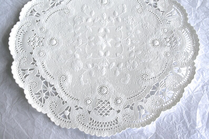 K&ouml;nnte beinhalten: Wei&szlig;e Papier-Deko-Serviette mit einem zarten Spitzenmuster. Die Serviette hat einen gewellten Rand und ein florales Design in der Mitte.