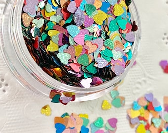 May include: A clear plastic pot filled with multi-coloured heart-shaped glitter. The glitter pieces are in various colours, including red, pink, yellow, green, and blue. Some hearts have a textured pattern. Glitter is scattered around the pot.
