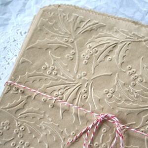 May include: A stack of brown paper gift bags with a holly leaf and berry pattern. The bags are tied together with red and white twine.