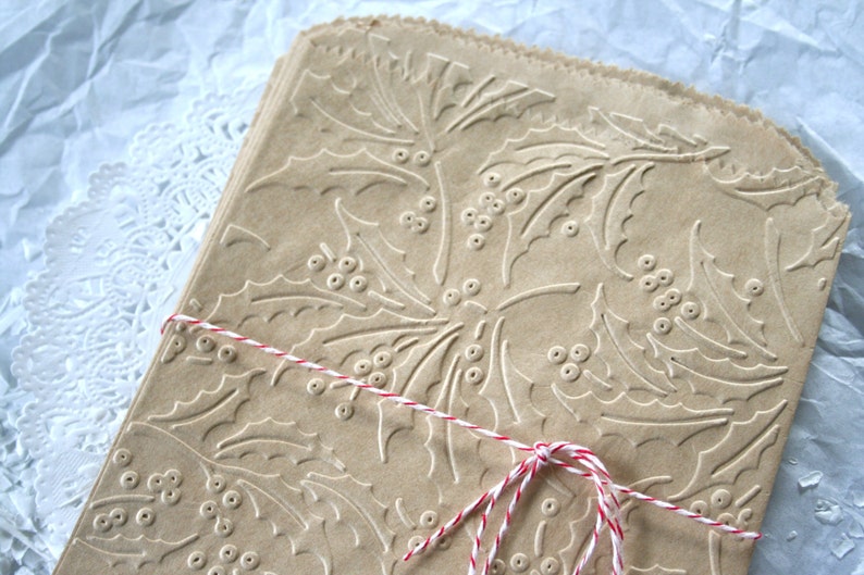 May include: A stack of brown paper gift bags with a holly leaf and berry pattern. The bags are tied together with red and white twine.
