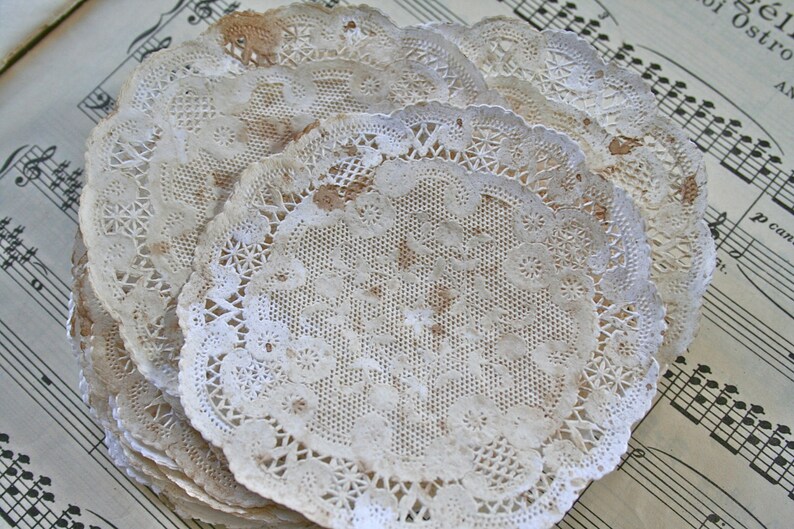May include: A stack of white paper doilies with a delicate lace pattern. The doilies are slightly worn and have a vintage aesthetic.