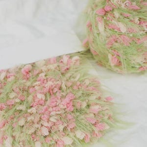 Pode incluir: Close-up de duas bolas de l&atilde;. Uma &eacute; uma bola, a outra tem formato retangular. Ambas s&atilde;o feitas de fio de l&atilde; verde, rosa e creme. O fio &eacute; macio e texturizado, com uma apar&ecirc;ncia delicada e artesanal. O fundo &eacute; branco.