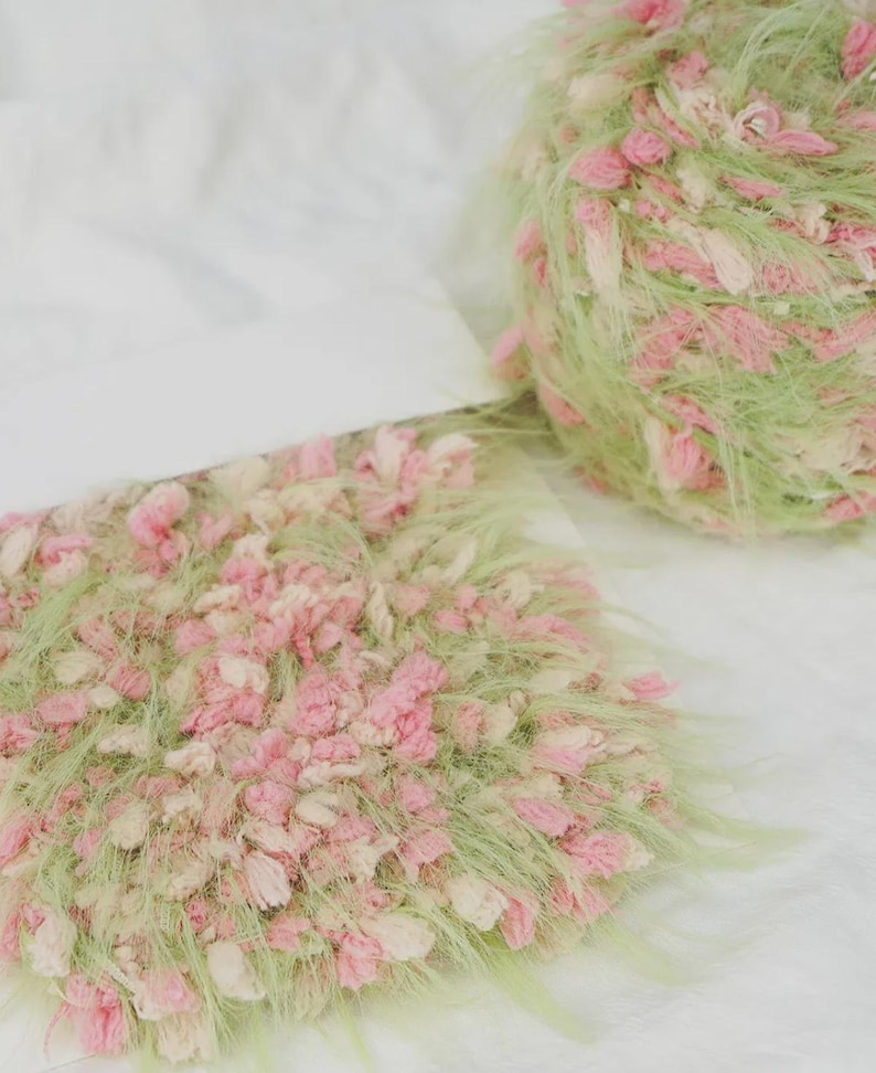 Pode incluir: Close-up de duas bolas de l&atilde;. Uma &eacute; uma bola, a outra tem formato retangular. Ambas s&atilde;o feitas de fio de l&atilde; verde, rosa e creme. O fio &eacute; macio e texturizado, com uma apar&ecirc;ncia delicada e artesanal. O fundo &eacute; branco.