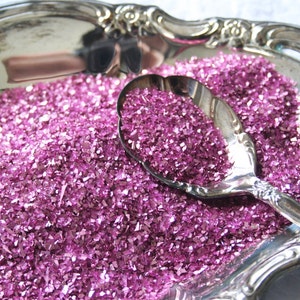 May include: A silver serving dish filled with pink iridescent glitter. A silver spoon is resting on top of the glitter.