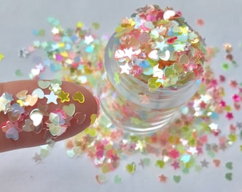 May include: A small glass jar filled with iridescent glitter in the shapes of hearts, stars, and other small shapes. The glitter is scattered on a white surface.