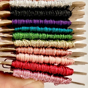 May include: A collection of 12 spools of metallic thread in various colors, including silver, black, purple, teal, green, gold, copper, pink, red, and rose gold. The spools are arranged in a stack and are held in a hand.