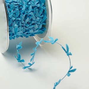 May include: A spool of light blue decorative ribbon with a textured, shredded appearance. The ribbon is made of a soft material and is arranged in a continuous strand, with small leaf-like accents. The ribbon is on a clear plastic spool.