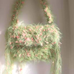Pode incluir: Uma bolsa fofa, feita &agrave; m&atilde;o, em verde claro, adornada com detalhes rosa. A bolsa apresenta uma al&ccedil;a de ombro e fios pendurados, criando uma apar&ecirc;ncia macia e texturizada. Um acess&oacute;rio &uacute;nico.