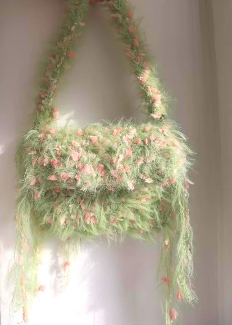 Pode incluir: Uma bolsa fofa, feita &agrave; m&atilde;o, em verde claro, adornada com detalhes rosa. A bolsa apresenta uma al&ccedil;a de ombro e fios pendurados, criando uma apar&ecirc;ncia macia e texturizada. Um acess&oacute;rio &uacute;nico.
