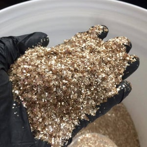 Peut inclure: Une main gant&eacute;e tient une grande quantit&eacute; de paillettes dor&eacute;es iris&eacute;es. Les paillettes sont compos&eacute;es de petites pi&egrave;ces plates qui r&eacute;fl&eacute;chissent la lumi&egrave;re.