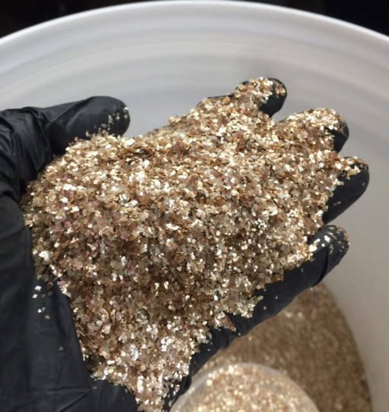 Peut inclure: Une main gant&eacute;e tient une grande quantit&eacute; de paillettes dor&eacute;es iris&eacute;es. Les paillettes sont compos&eacute;es de petites pi&egrave;ces plates qui r&eacute;fl&eacute;chissent la lumi&egrave;re.