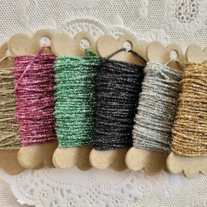 May include: Six spools of sparkly thread in gold, pink, green, black, silver, and gold. The thread is wound around cardboard spools with a heart shape.