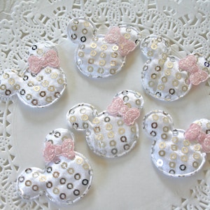 May include: Six white fabric mouse ears with pink bows and gold sequins. The mouse ears are on a white doily.