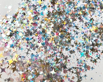 May include: A close-up of iridescent glitter in various shapes, including stars, scattered on a white background.