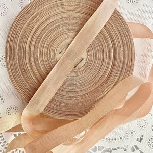 May include: A spool of beige ribbon with a satin finish. The ribbon is wound around a cardboard spool and is lying on a white lace surface.