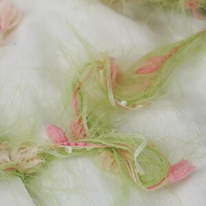 Pode incluir: Close-up de um tecido com uma apar&ecirc;ncia fofa e texturizada. O tecido apresenta fios de fios verde claro, rosa e branco, criando um efeito macio e em camadas. O design inclui pequenos detalhes rosa em forma de flor.