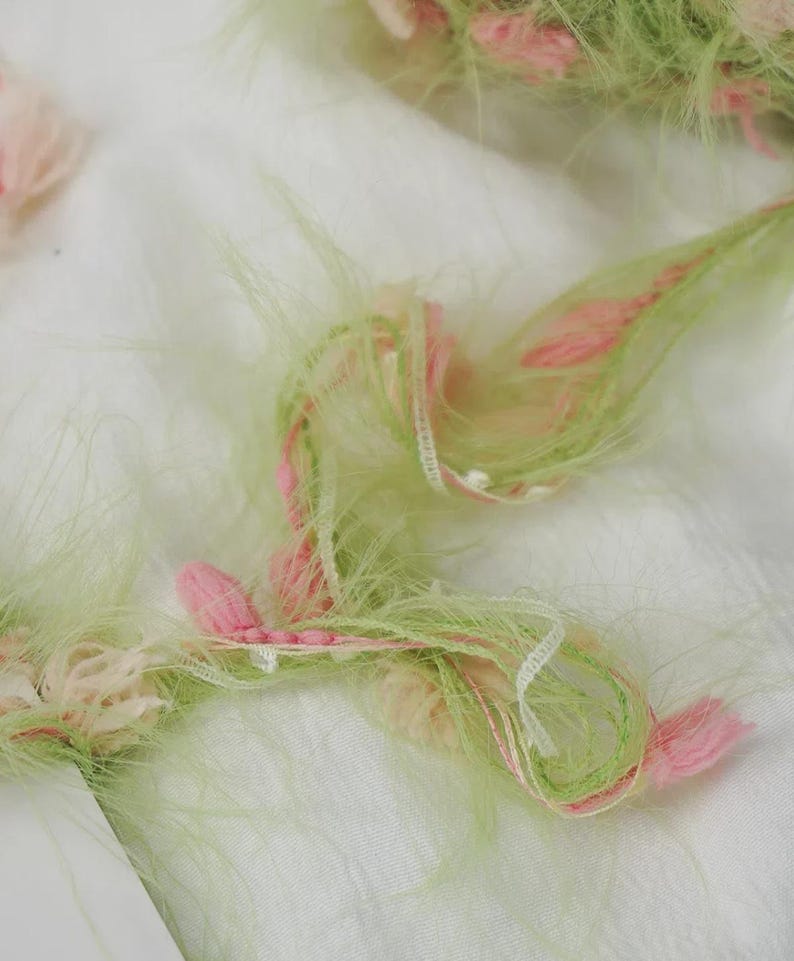 Pode incluir: Close-up de um tecido com uma apar&ecirc;ncia fofa e texturizada. O tecido apresenta fios de fios verde claro, rosa e branco, criando um efeito macio e em camadas. O design inclui pequenos detalhes rosa em forma de flor.